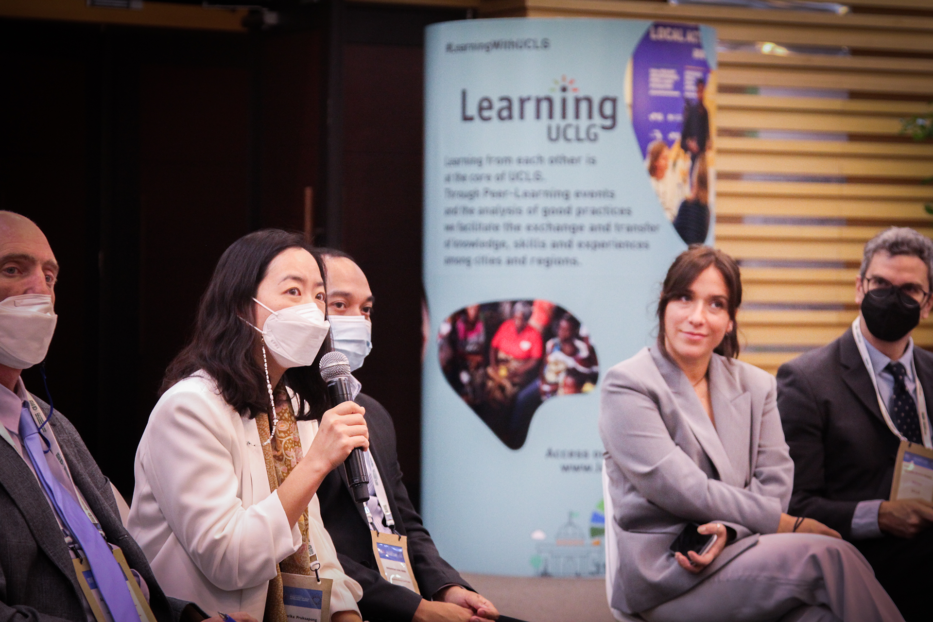 1.	A woman wearing a face mask holds a mic while sitting in between other persons. A column with UCLG Learning’s identity shows in the background.